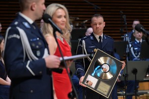 Na zdjęciu widać policjanta trzymającego złotą płytę oraz kobietę i policjanta.
